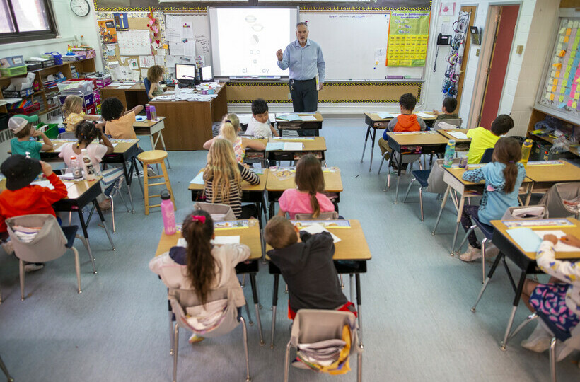 Teacher stands at front of the room and instructs students.